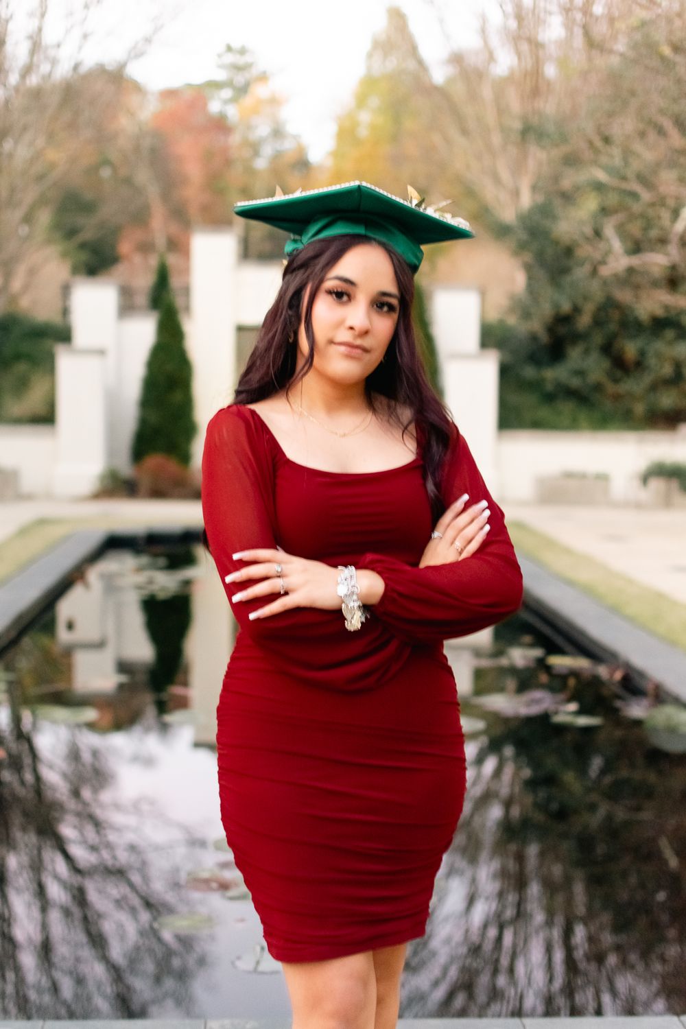 Graduate in burgundy dress and green cap poses confidently outdoors.