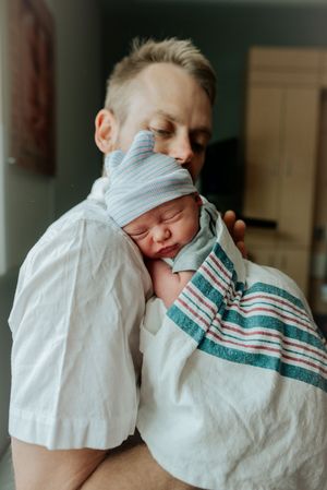 Father gently kissing sleeping newborn baby wrapped in striped hospital blanket.