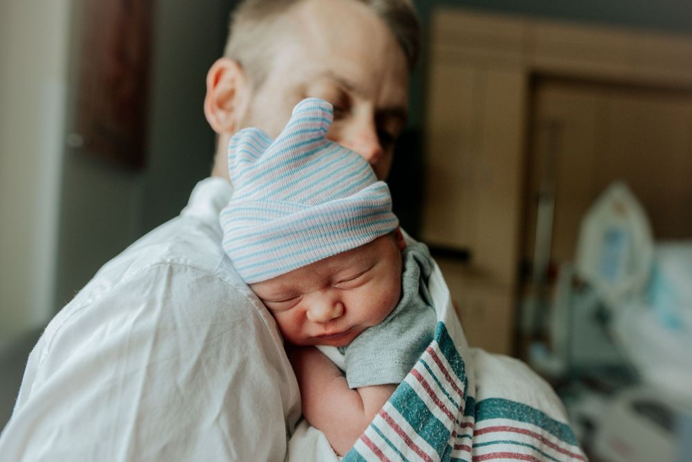 Close-up of father cradling sleeping newborn in striped blanket wearing hospital hat.