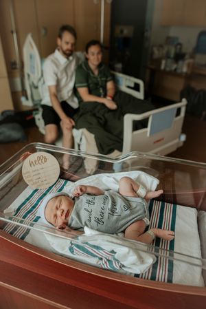 Newborn baby in hospital bassinet with proud parents resting on bed in background.