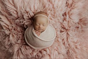 Sleeping newborn baby girl wrapped in pink knit swaddle with floral headband on a soft pink fluffy rug.