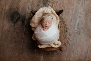 Newborn baby sleeping peacefully wrapped in white knit swaddle with cream bonnet inside a rustic wooden bowl. Ivy Lynn Photography