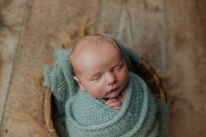 Peaceful newborn baby swaddled in a soft teal knit wrap resting in a wicker basket.