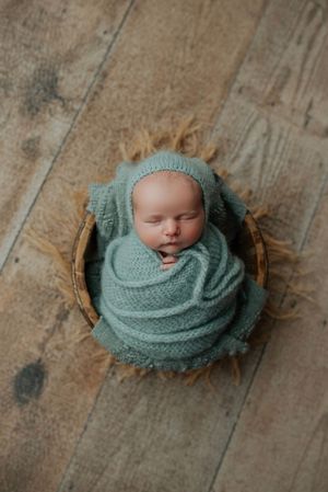 Newborn baby wrapped in teal knit fabric with matching bonnet sleeping in a wicker basket.