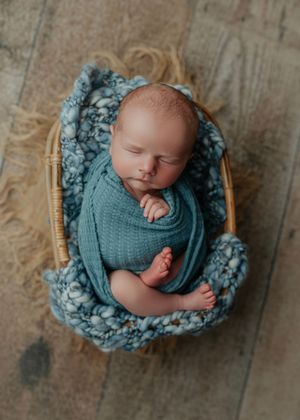 Sleeping newborn swaddled in blue wrap with tiny feet peeking out in a wicker basket.