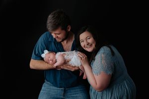 Smiling parents cradling sleeping newborn baby in white wrap against black background.