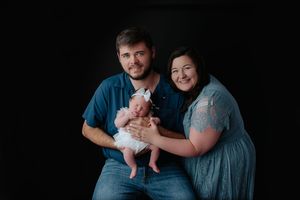 Happy couple holding newborn baby in white outfit against dark studio background.