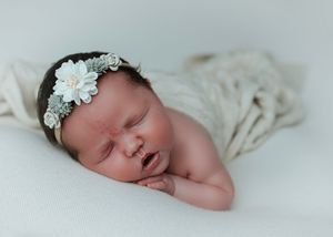 Sleeping newborn baby girl wearing floral headband wrapped in cream blanket.