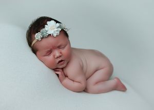 Peaceful newborn baby girl with floral headband posed on soft white backdrop.