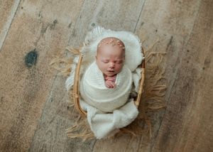 Newborn baby wrapped in cream knit swaddle with pearl headband sleeping in wicker basket.