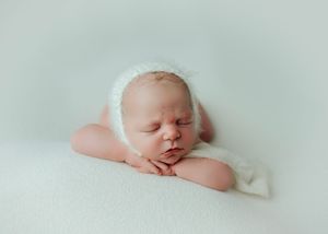Sleeping newborn wearing a fluffy white bonnet posed on a soft white blanket.