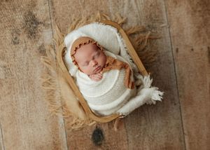Sleeping newborn swaddled in cream wrap with brown bonnet resting in wooden basket on wood floor.