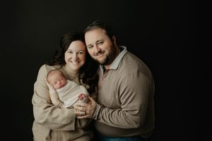 Happy new parents smiling and holding their swaddled newborn baby against a dark black studio background. Ivy Lynn Photography