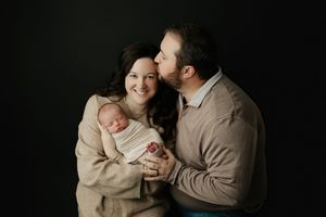 Loving father kissing mother on cheek while she holds their swaddled newborn baby in a dark studio setting. Ivy Lynn Photography