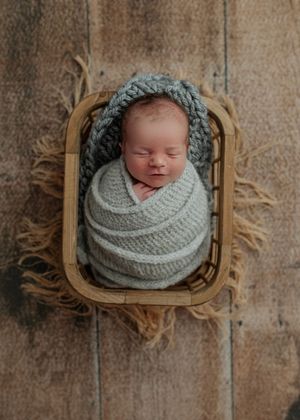 Sleeping newborn swaddled in gray knit wrap with matching bonnet resting in rustic wooden basket.