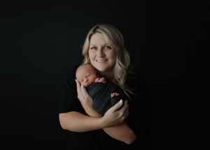 Blonde woman smiling while holding swaddled newborn baby against dark black background.