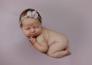 Sleeping newborn baby girl posed on mauve backdrop wearing a delicate pink bow headband.