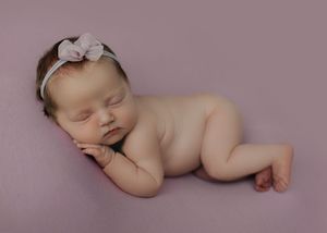 Peaceful newborn baby girl in froggy pose on purple backdrop with pink bow headband.