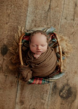 Sleeping newborn baby swaddled in brown wrap and bonnet in wicker basket prop.