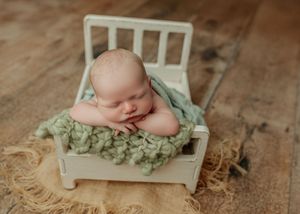 Peaceful newborn baby posed on tiny white bed prop with green knit blanket.