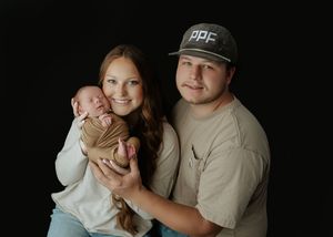 Young couple smiling and holding their swaddled newborn baby against a solid black studio background.