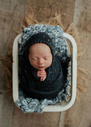 Newborn baby wrapped in dark navy swaddle with matching knit bonnet sleeping in a white wicker basket.