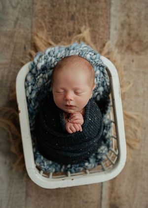 Peacefully sleeping newborn swaddled in dark navy wrap resting in a white wicker basket on wood floor.