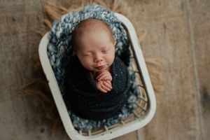 Sleeping newborn baby wrapped in dark blue knit blanket inside a small white basket with blue textured lining.