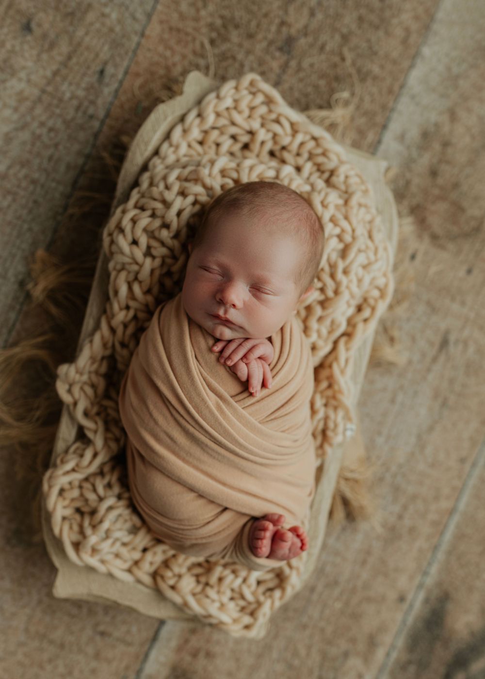 Peacefully sleeping newborn wrapped snugly in a warm beige swaddle atop a chunky knit blanket in a wooden bowl.