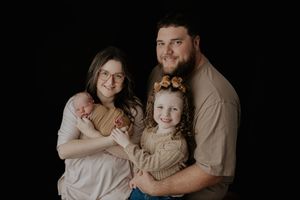 Family portrait of parents with newborn baby and smiling toddler girl against black backdrop.