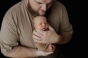 Bearded father tenderly kissing his sleeping newborn baby wrapped in a tan swaddle against a dark background.