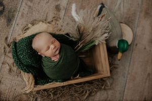 Sleeping newborn in green knit hat and wrap nestled in a wooden crate with hunting and outdoor props.