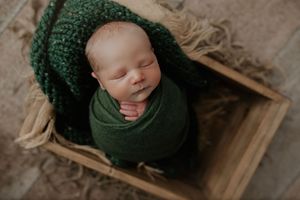 Sleeping newborn baby swaddled in green wrap with matching knit hat nestled in a rustic wooden crate.