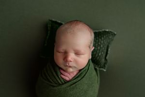 Peacefully sleeping newborn wrapped snugly in a green swaddle on a matching green backdrop with small pillow.