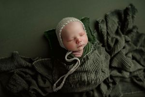 Newborn baby in cream knit bonnet swaddled in green wrap resting on textured green blanket backdrop.