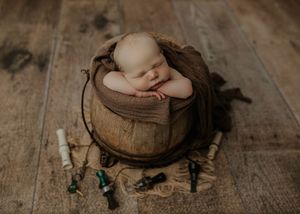 Sleeping newborn posed in a rustic wooden bucket with brown wrap surrounded by fishing lures on wood floor.