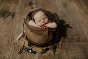 Newborn baby sleeping peacefully in a wooden bucket with brown wrap and fishing lures on rustic wood floor.