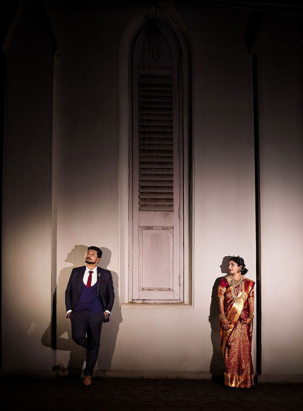A couple in traditional wedding attire poses dramatically against a white wall with arched shutters.