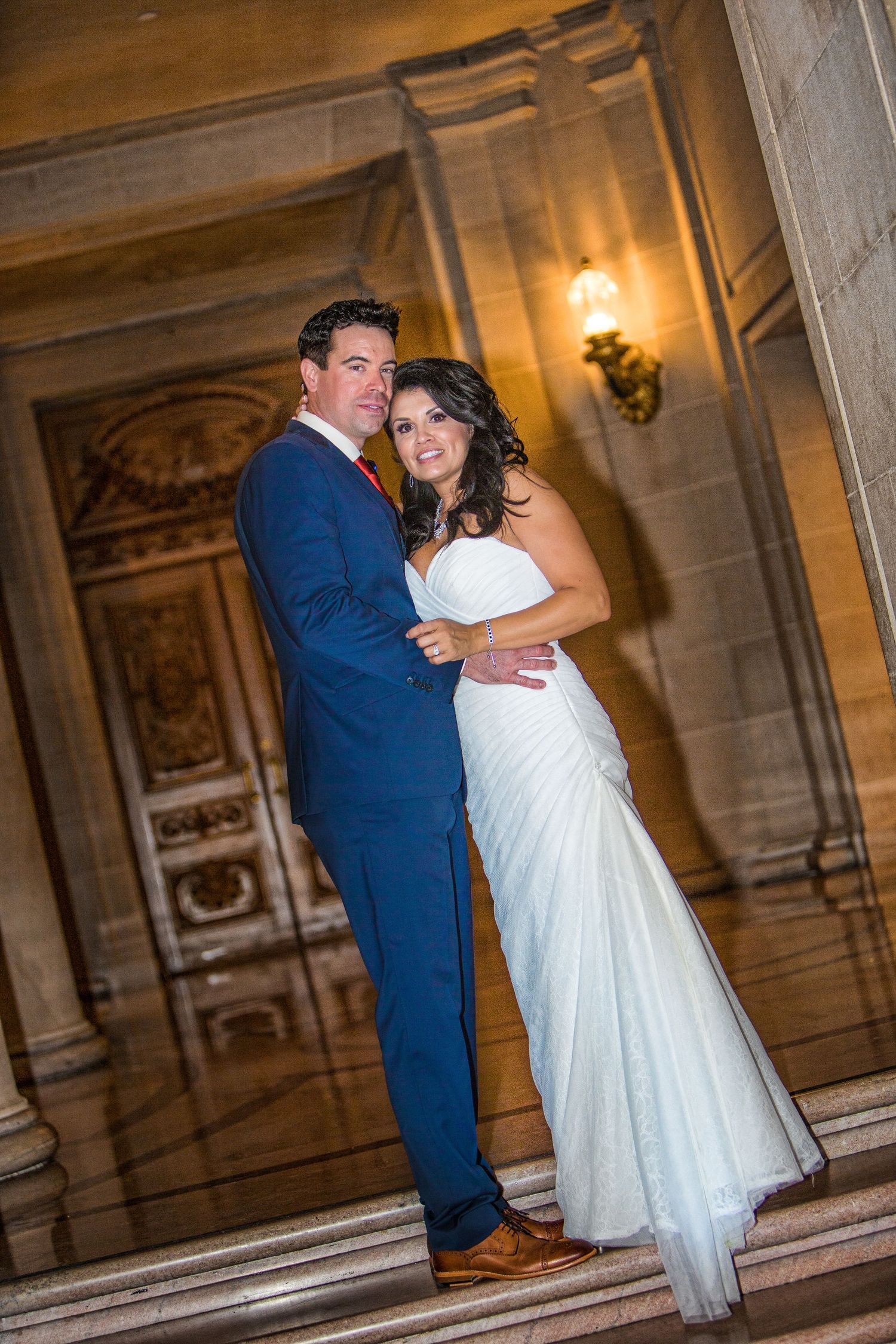 A bride in a white strapless gown smiles with her groom in a dark suit under romantic lighting at their wedding reception.