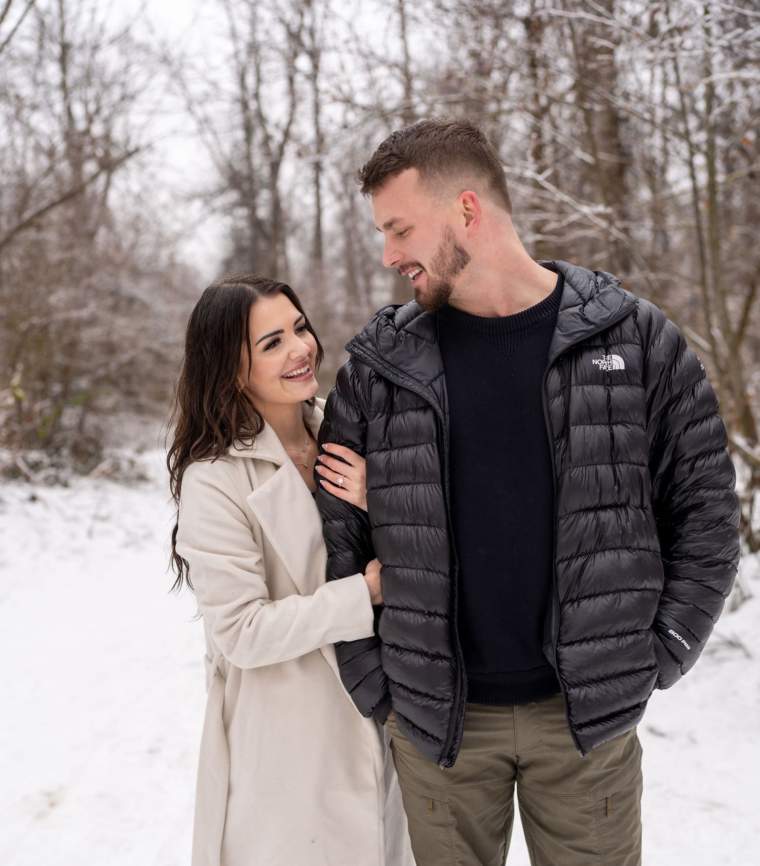 A couple dressed in black walking together by a snow-covered lake with mountains in the background.