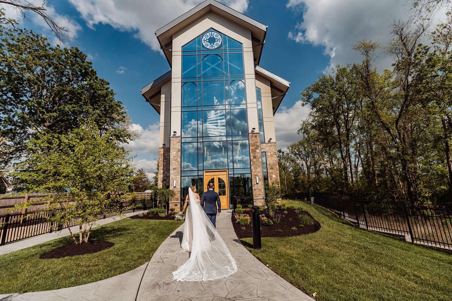 Bride and groom pose in front of a modern glass chapel with brick accents surrounded by lush greenery on a sunny day.