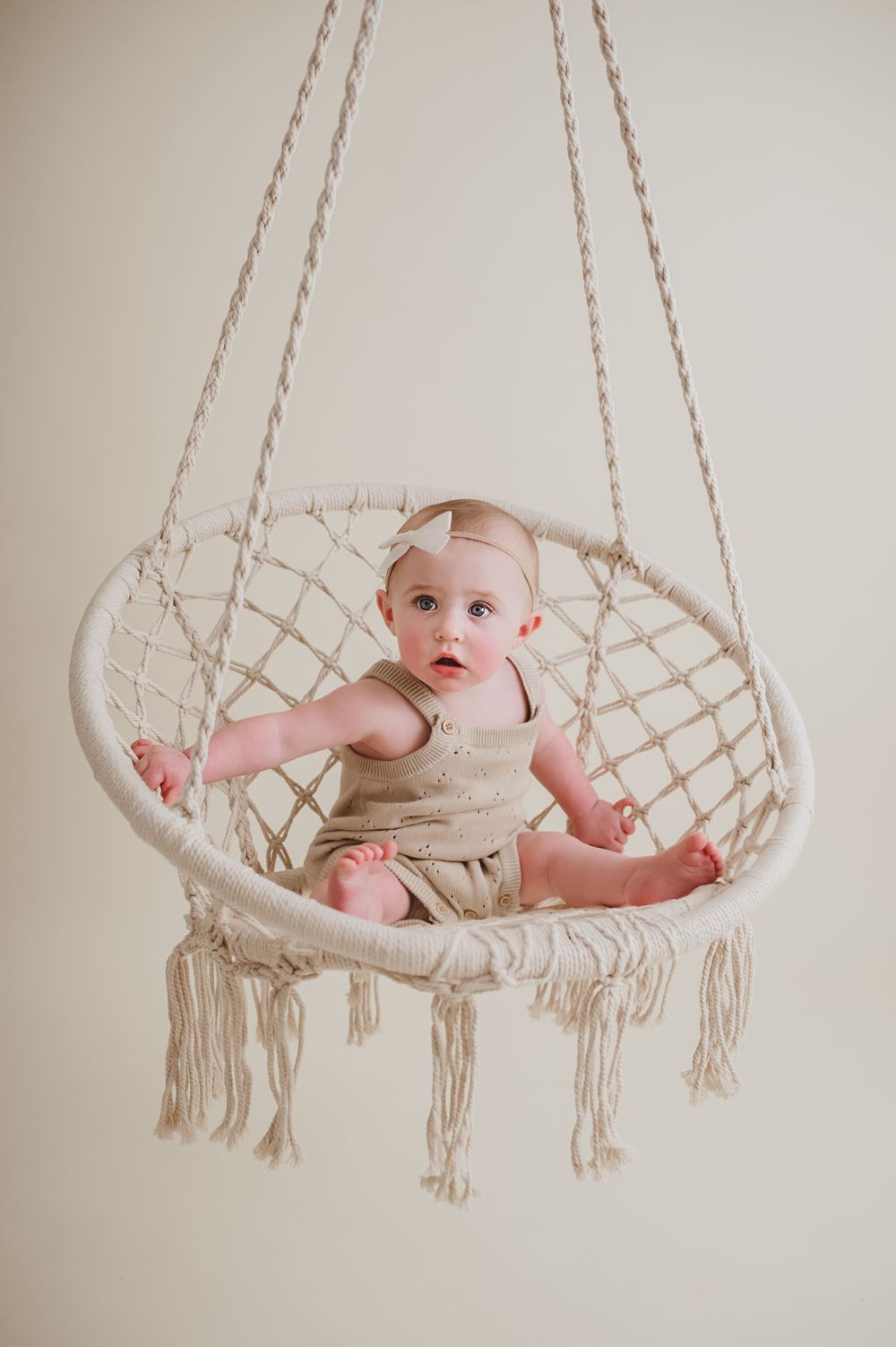 A macrame hanging chair with decorative tassels holds a small baby wearing neutral linen clothing against a beige backdrop for studio photos