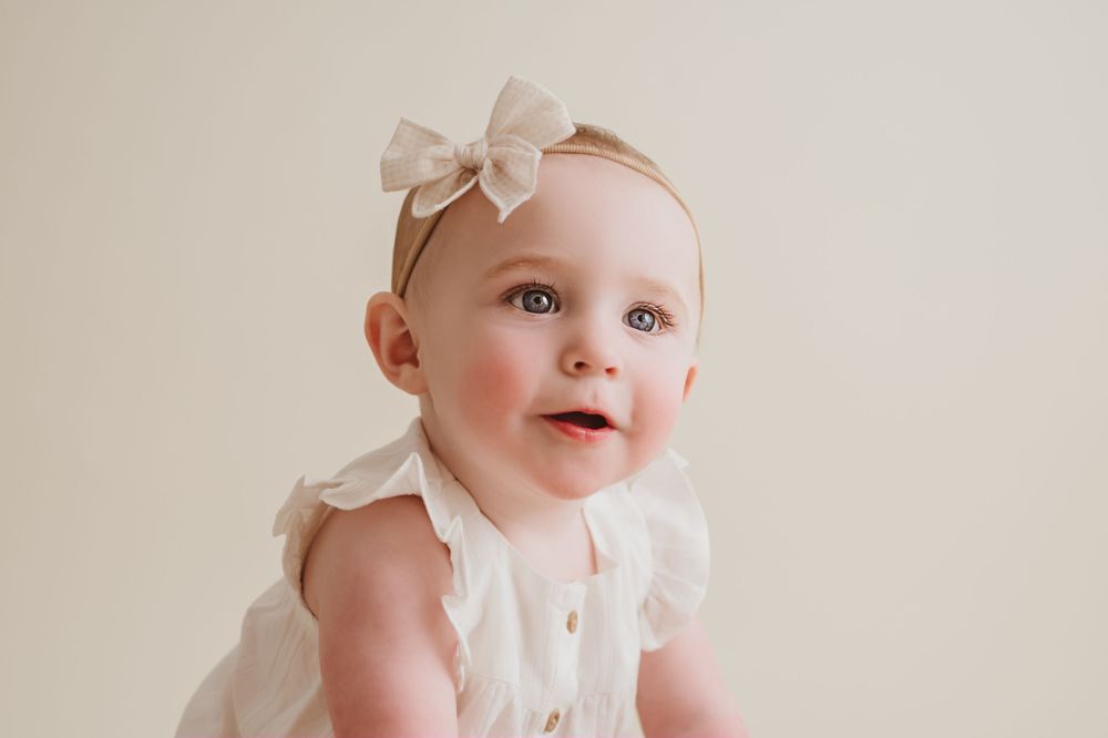 A smiling baby in white with cream bow headband against a neutral background for studio milestone session in Chattanooga