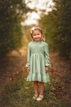 A small child in a mint green dress stands on a path through an apple orchard surrounded by autumn leaves at golden hour.