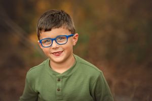 A young boy in blue glasses and green shirt stands outdoors against an autumn background.