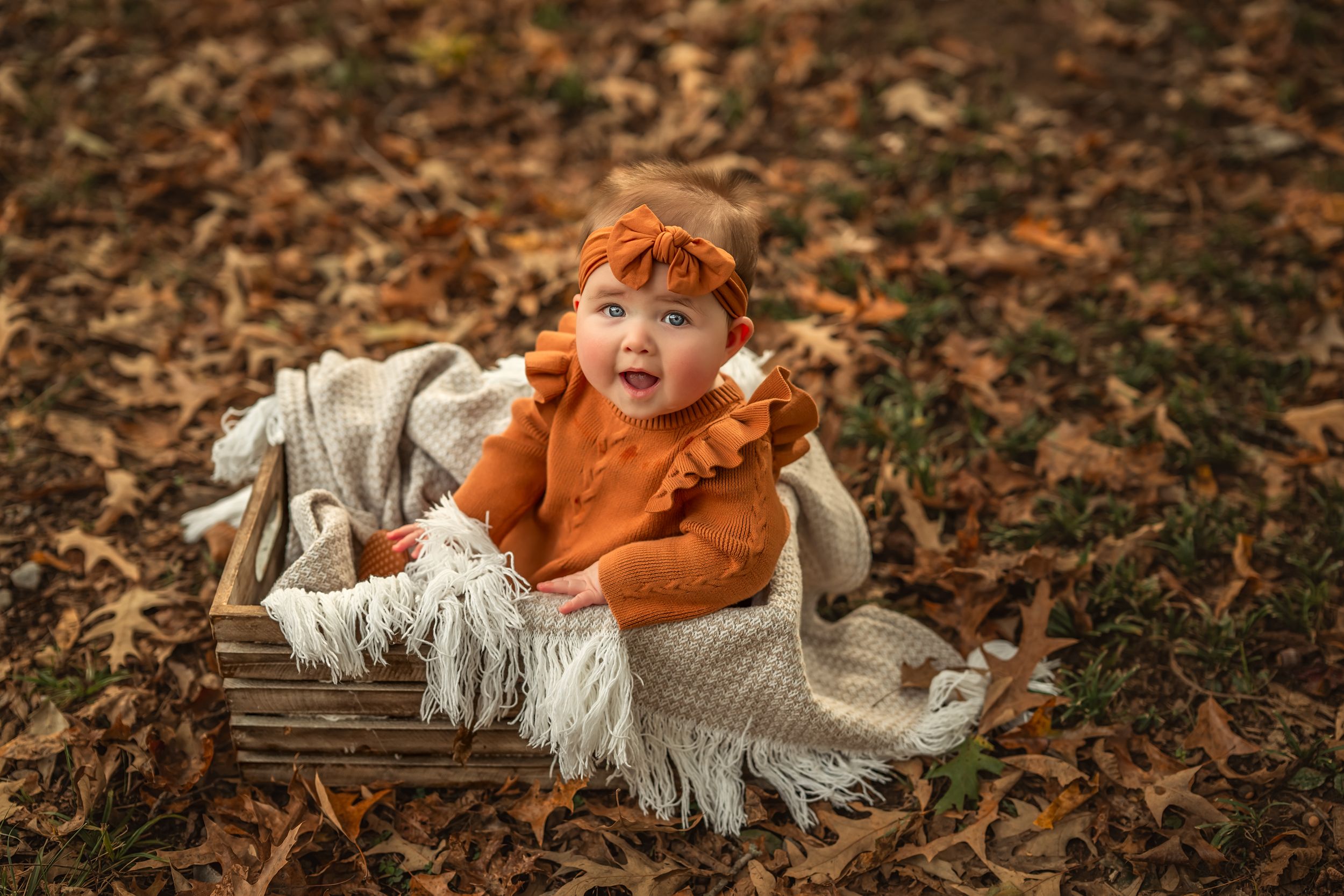 A baby in orange sits in a crate with a cozy blanket surrounded by scattered autumn leaves on the ground for milestone photos.