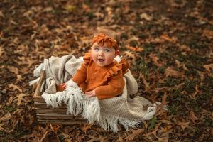 A baby in orange sits in a crate with a cozy blanket surrounded by scattered autumn leaves on the ground for milestone photos.