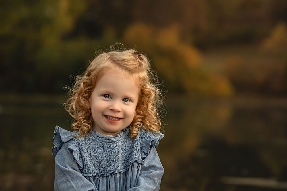 Young child with curly red hair wearing a ruffled denim dress smiles warmly outdoors during autumn.