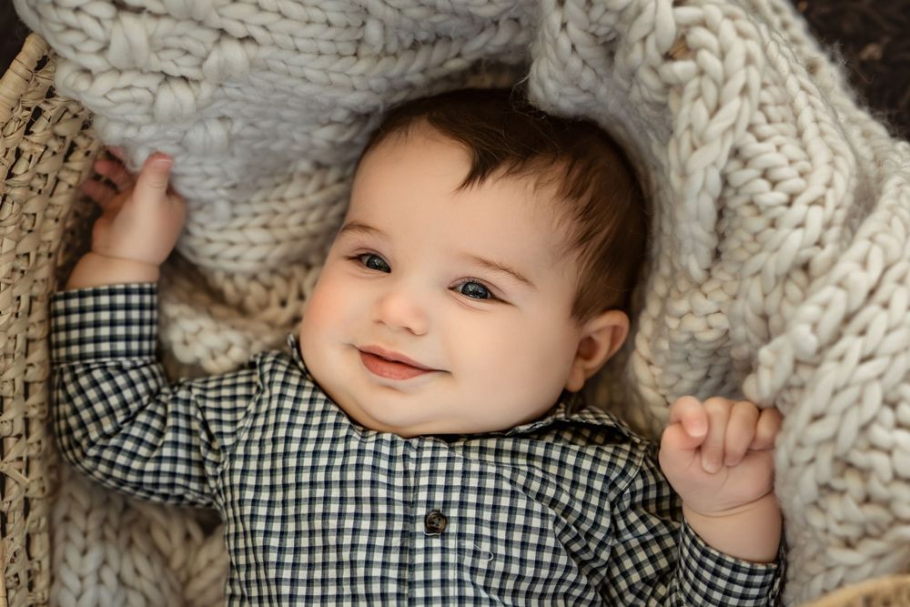 A smiling baby wearing a checkered shirt lies on a cozy cream knit blanket.