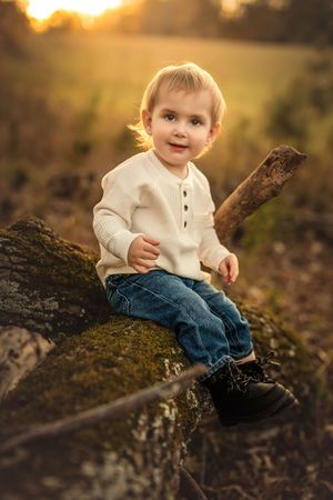 An 18-month-old boy in jeans and white sweater sits on a mossy log at sunset for milestone session with Atlanta Children's photographer.